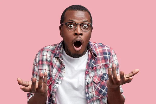 Frustrated African American male feels puzzled and hesitant, shrugs shoulders in bewilderment, keeps mouth widely opened, doesn`t know what to do, isolated over pink background.