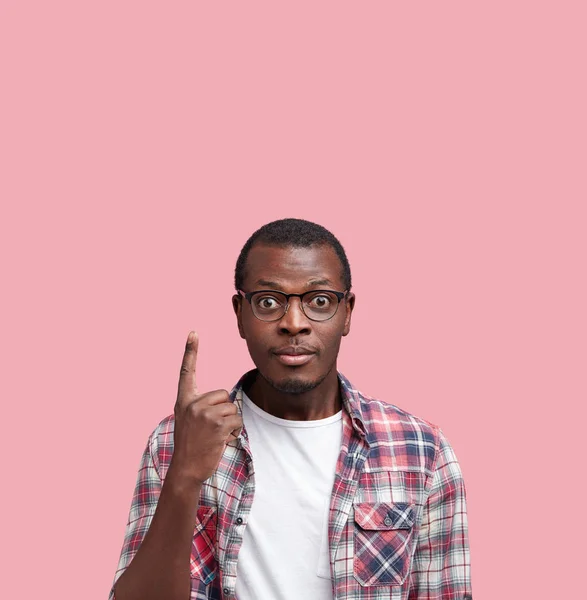 Vertical shot of confused handsome African American male with dark healthy skin, wears shirt and spectacles, poses alone at blank copy space, advertises something. Look there!