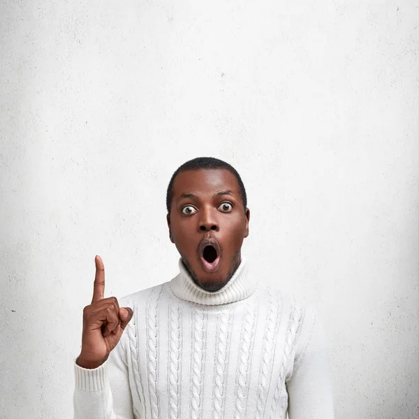 Shocked bugged eyed dark skinned man with surprised expression, wears white sweater, indicates with fore finger upwards, demonstrates blank copy space for your advertising content or promotion
