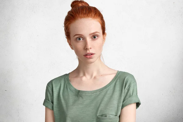 Lovely freckled woman with ginger hair knot, dressed casually, listens attentively lecture, isolated over white background. Serious ginger young female looks mysteriously at camera, poses indoor