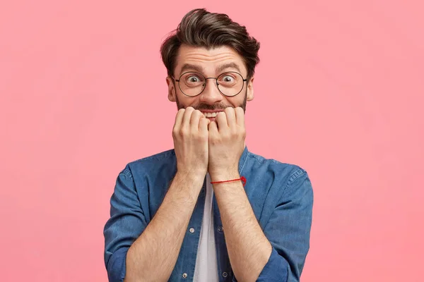 Portrait of nervous male bites finger nails, has trendy hairdo, wears denim casual shirt, looks worried and excited before important event, isolated over pink studio background.