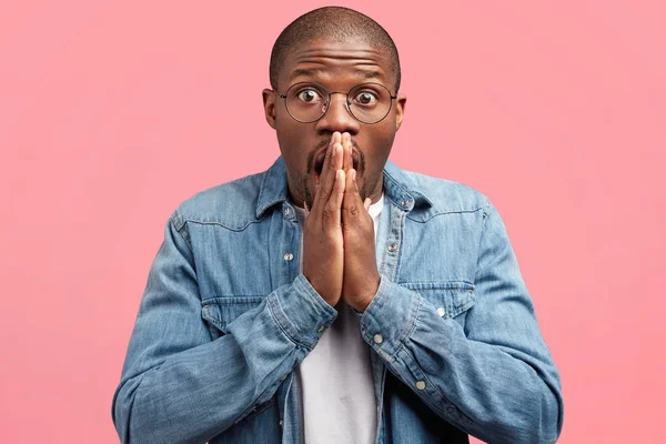 Headshot of emotional frightened dark skinned male keeps palms pressed together, looks with frustrated expression, hears shocking news on television, poses against pink background.