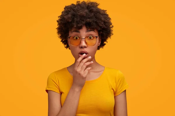 Horizontal shot of surprised African American female in bewilderment, feels shocked, keeps mouth opened, listens to exciting story, isolated over yellow background.