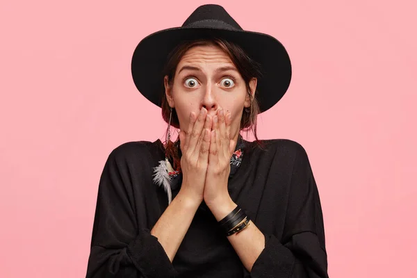 Horizontal shot of frightened young Caucasian female in stylish black hat, stares at camera with full disbelief and scared expression, shocked with unexpected news, isolated on pink background