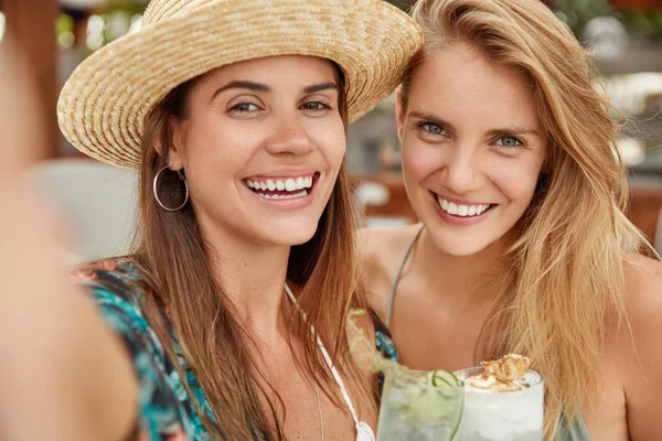 Photo of delighted females with broad smiles, pose together for making selfie, enjoy good recreation, drink summer beverages or cocktails, being in good mood.