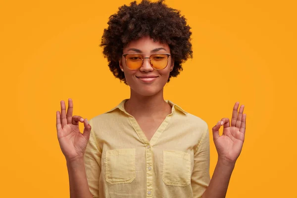 Positive dark skinned woman with cheerful expression, makes ok sign, wears elegant trendy shades and shirt, being satisfied, isolated over yellow background.