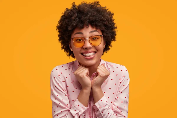 Horizontal shot of happy dark skinned young female with positive expression, being touched by receiving compliment about her appearance, keeps hands together, isolated over yellow background