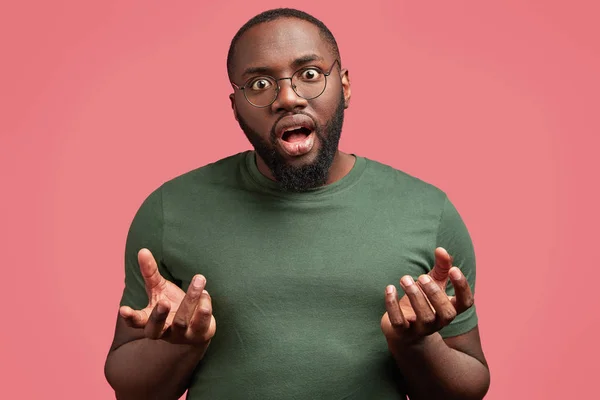 Puzzled bearded African American male gestures with uncertainty and looks in bewilderment, being astonished by unexpected news, stands against pink studio background. People and facial expressions