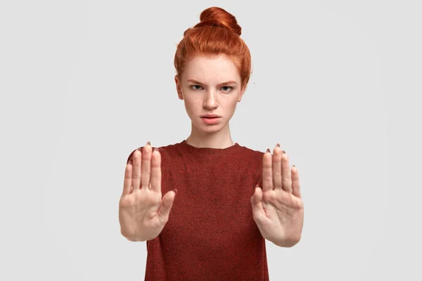 Leave me in peace, please! Don`t bother! Angry red haired female pulls palms at camera, shows stop gesture, wears casual clothing, isolated on white background.