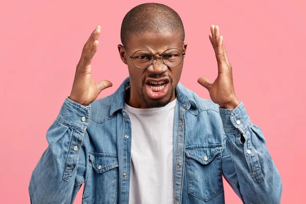 People, negative emotions and reaction concept. Irritated African American guy has big troubles, gestures angrily with hands, frowns face in dissatisfaction, wears casual denim shirt, isolated on pink