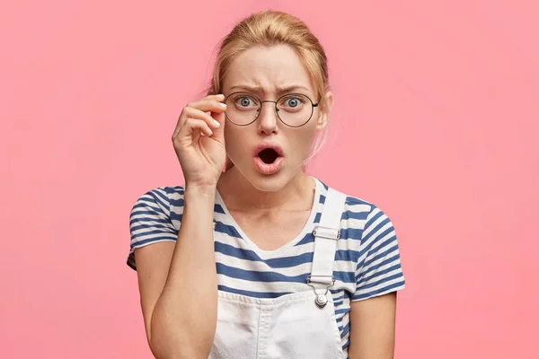 Scared beautiful blonde female with amazed expression, keeps hand on eyewear, keeps mouth wide opened, dressed in striped t shirt, stands against pink background.