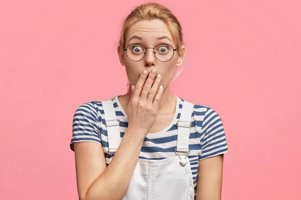 Terrified female with blonde hair and blue eyes, covers mouth with hand, has worried expression, dressed in casual clothing, stares through spectacles, being puzzled by bad news about health