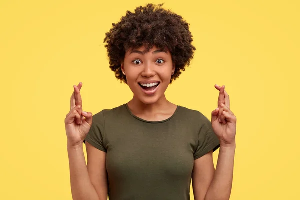 Photo of lovely cheerful African American woman in high spirit, crosses fingers as has big hope in something, makes desirable gesture, stands against yellow background.