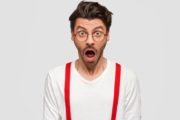 Emotive bearded male with trendy hairstyle, keeps jaw dropped, has shocked face, isolated over white background. Surprised hipster with eyes popped out