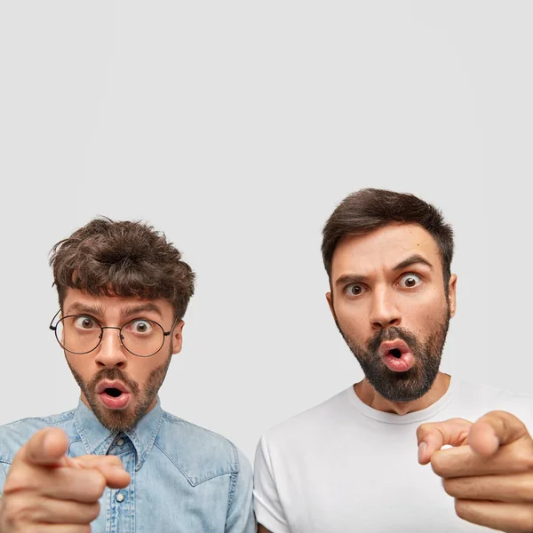 Surprised comic men point at camera with stunned expressions, see something unbelievable, stand close to each other, frown faces from shock, isolated over white background.