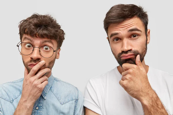 Confused two male colleagues frown faces with doubt, think over how overcome financial problems in company, have trendy haircuts, hold chins, pose against white blank background.
