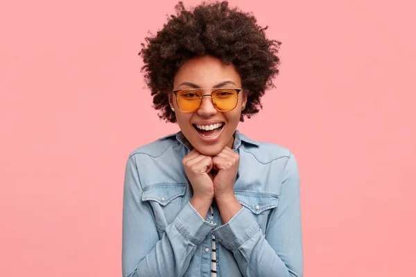 Headshot of beautiful African American female keeps hands under chin from happiness, smiles positively, rejoices good proposal from partner, wears jean fashionable clothes.