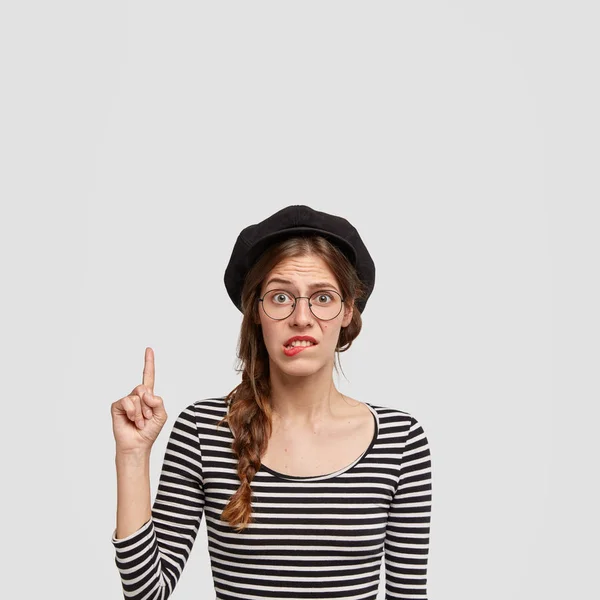 Hesitant Parisian female bites lower lip in puzzlement, points upwards with index finger, feels clueless whether to buy something, dressed in striped sweater, isolated over white background.