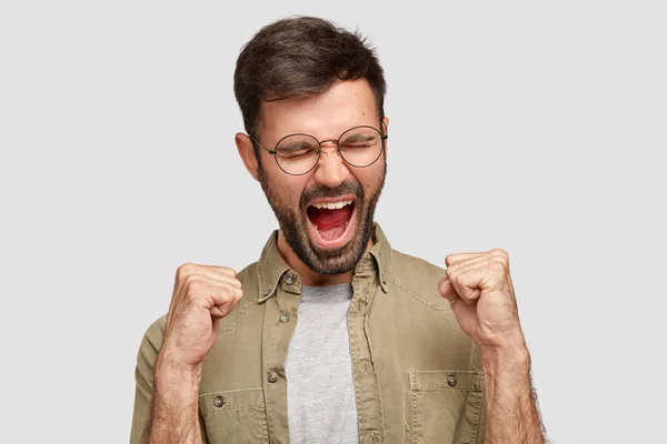 Crazy guy clenches fists and shouts angrily, expresses aggression and discontent, frowns face, feels wicked, dressed in fashionable shirt, stands against white background.