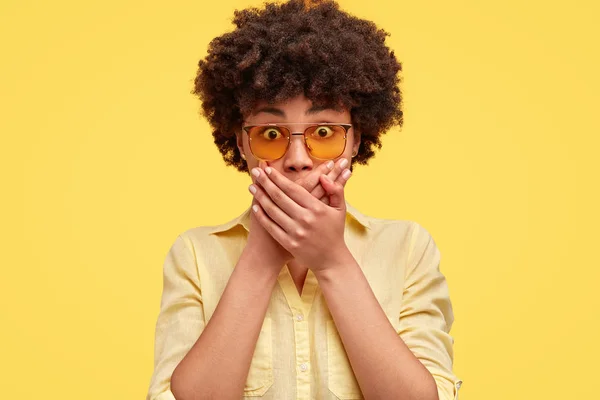 Dark skinned young female has scared expression, covers mouth with hands, looks in astonishment, wears trendy shades and blouse, expresses great surprise, isolated over yellow background.