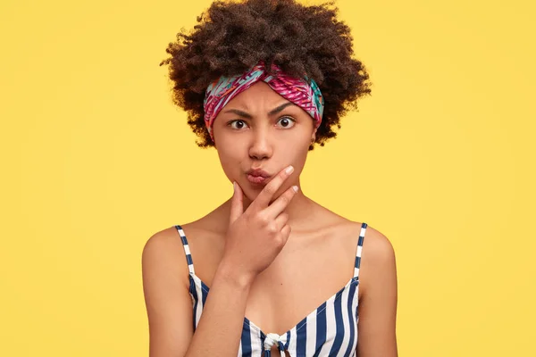 Photo of serious displeased female with Afro curly hairstyle, holds chin, frowns face, dressed in striped clothes, has strict expression, isolated over yellow background.