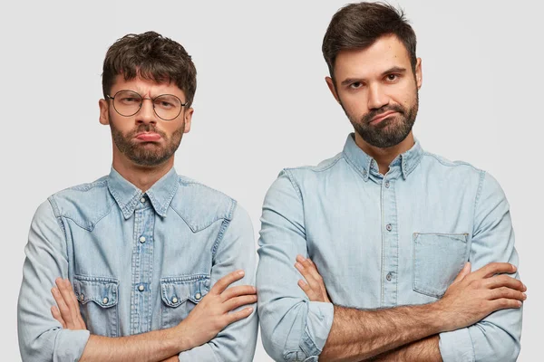 Two displeased guys keep arms folded, look with sullen expression, feel like loosers after loosing game, dressed in fashionable denim clothes, stand next to each other over white background.