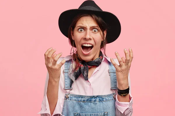 Horizontal shot of beautiful annoyed female agricultural worker gestures, has irritated facial expression, being displeased with work, wears black hat and denim dungarees isolated over pink background