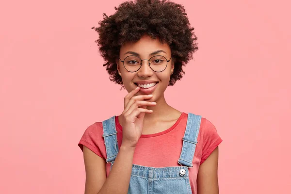 Glad dark skinned curly female with pleasant smile, dressed in casual t shirt with denim overalls, isolated over pink background. Beautiful mixed race student rejoices successfully passed exam.