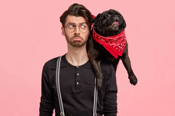 Displeased bearded male purses lips, looks with negative facial expression at his dog, being discontent after walk, pose together against pink background.