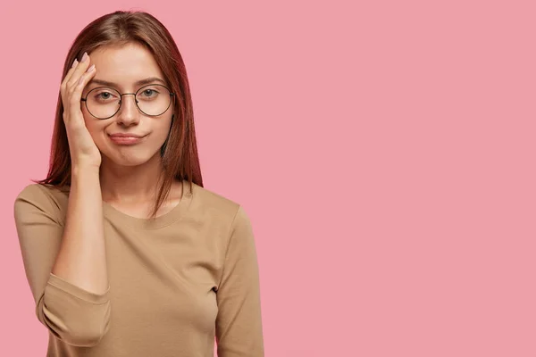 Horizontal shot of lovely female has bored displeased facial expression, looks in discontent, wears round spectacles and casual sweater, isolated over pink background with blank copy space aside