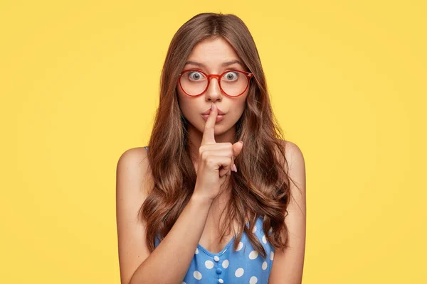 Horizontal shot of attractive young female shows silence sign, asks to be close mouthed and speechless, has shocked expression, wears trendy outfit, spectacles, isolated over yellow background