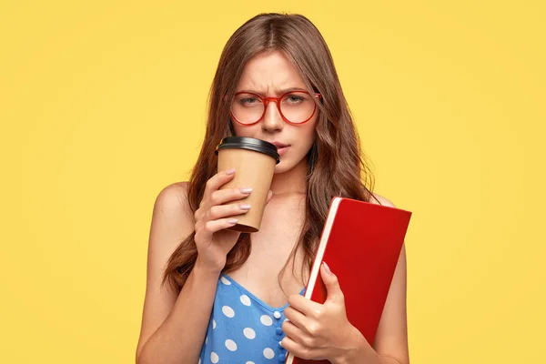 Low spirited young female student feels sad after receiving bad mark for exam, being tired, drinks hot coffee, holds scientific literature, stands against yellow background.