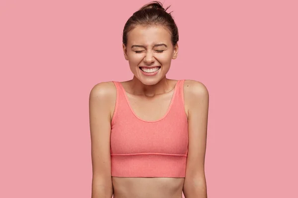 Positive female in sports top, giggles happily, likes gymnastics, closes eyes from happiness, glad to achieve great results in sport, poses against pink background.