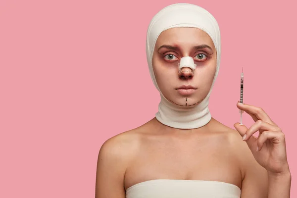 Photo of woman has marked face with lines, bruised skin, holds syringe for making facial rejuvenation procedure, undergoes plastic surgery or cosmetic operation in clinic, isolated on pink wall