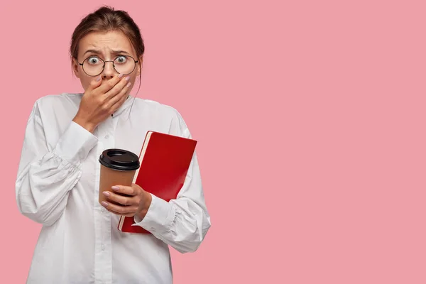 Terrified student girl covers mouth with shocked expression, stares with bugged eyes, holds takeaway coffee, red diary, receives negative results of exam, isolated over pink background. Omg concept