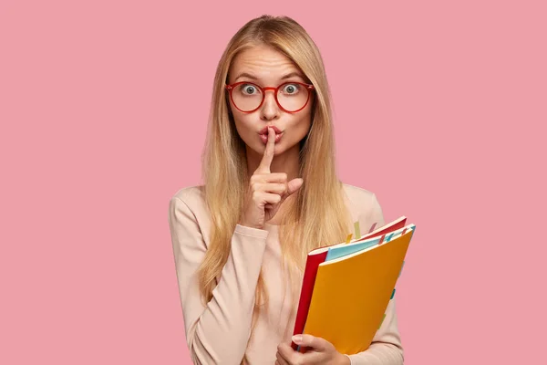 Omg, don\'t speak! Attractive Caucasian girl keeps index finger over lips, demonstrates silence gesture, asks not bother her, be quiet and speechless, wears spectacles with red rim isolated on pink wall