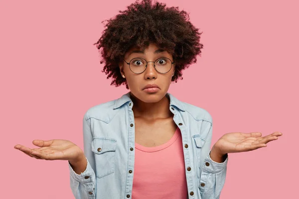 Doubtful dark skinned girl makes scale with arms, feels hesitant and puzzled, finds answer to difficult question, dressed in fashionable outfit stands against pink background.