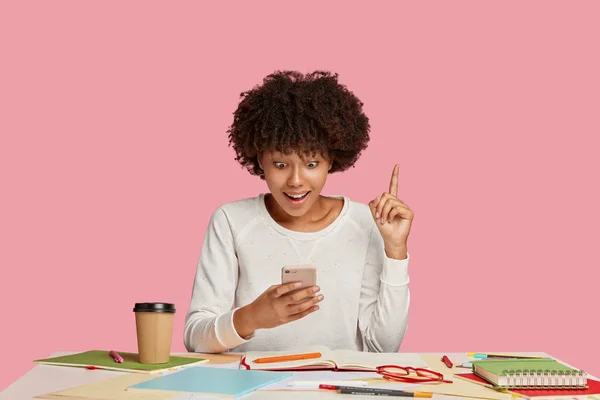 Horizontal shot of surprised happy woman blogger with Afro hairstyle, stares at cell phone, raises index finger, gets nice idea for developing website, works in cozy place, uses notepad and pen
