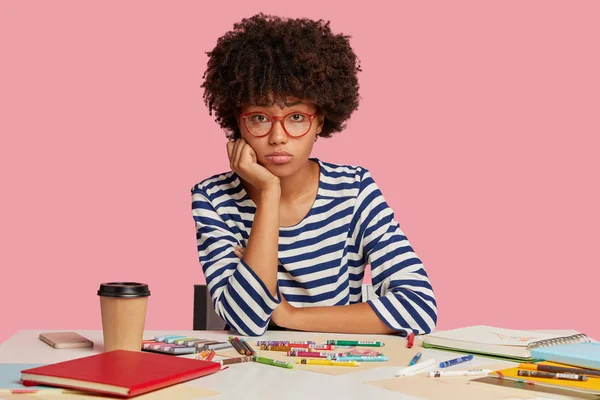 Sad woman painter with Afro haircut, has frustrated look at camera, doesn\'t have inspiration for creating masterpiece, surrounded with colorful markers, disposable cup of coffee, notepad with sketches