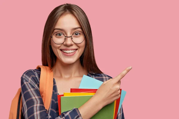 Horizontal shot of pleasant looking cheerful European girl points with index finger at blank copy space, advertises free space for your advertisement, carries textbooks and vocabularies, isolated