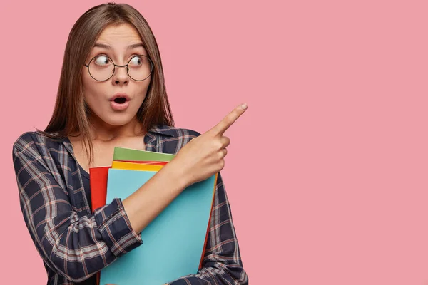 Horizontal portrait of attractive schoolgirl with surprised expression, points with index finger at blank copy space, advertises something, wears casual clothes, holds pile of books, isolated on pink