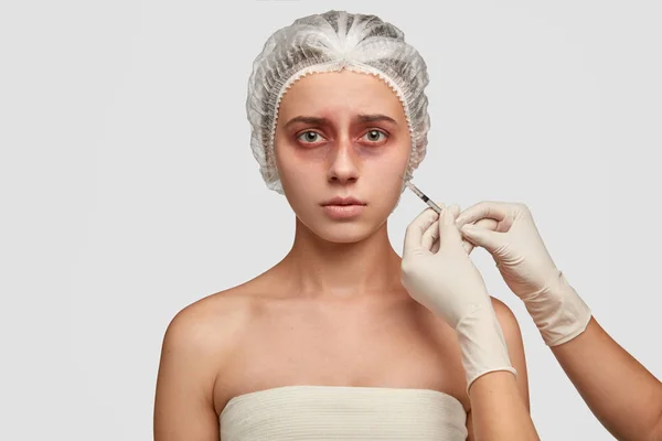 Horizontal shot of desperate bruised young woman receives botox injection, wants to change her appearance, has puzzled after plastic surgery, poses against white background.