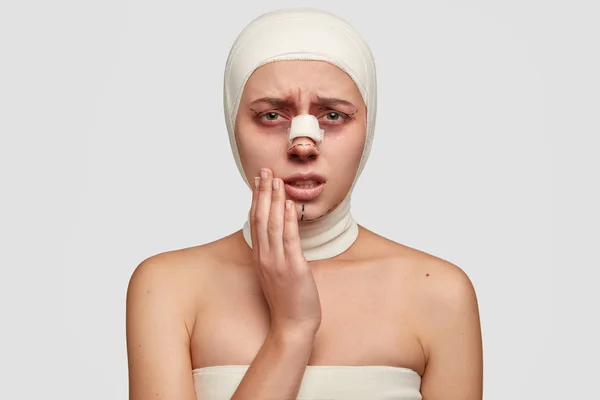 Desperate sick woman keeps hand on cheek, suffers from strong pain after skin grafting, has bandage or plaster on nose, isolated over white background.