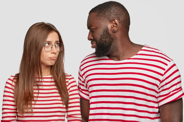 Photo of mixed race couple sort out relationships, look in displeasure at each other, have quarrel, dressed in similar t shirts, stand against white background.