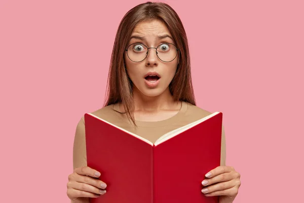 Stunned surprised teacher with book in hands, looks at pupil who made mess in classroom, wears transparent glasses, keeps jaw dropped, poses against pink background.