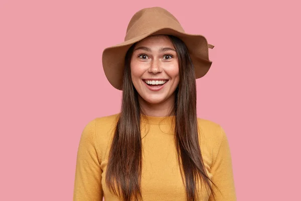 Joyful woman in romantic mood gazes with pleasant smile, rejoices relationships, wears fashionable hat and yellow clothes, isolated over pink background.