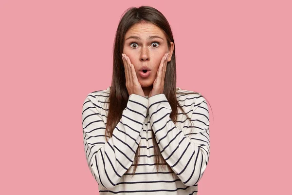 Shocked woman with terrified expression, keeps both hands on cheeks, gasps from wonder, dressed in casual striped sweater, poses over pink background.