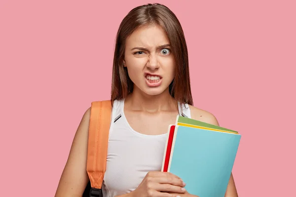 Image of annoyed dark haired schoolgirl frowns face and clenches teeth in discontent, feels peevish with bored classmate, tries to explain material, carries backpack and books poses on pink background