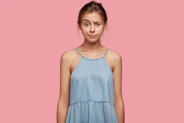 Indignant female youngster raises eyebrows with bewilderment, has unexpected reaction, wears casual jean summer dress, poses against pink studio background.