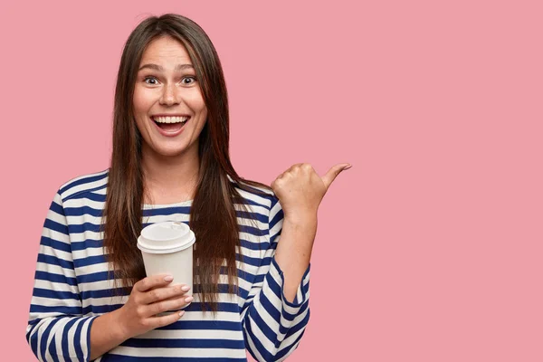 Indoor shot of pleasant looking positive woman with satisfied expression, spends free time with friends in coffee shop, holds disposable cup of beverage, dressed casually.
